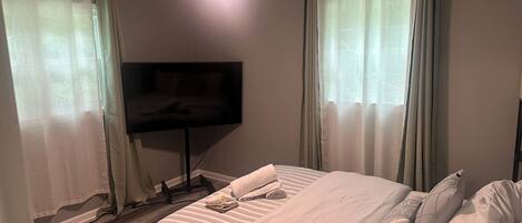 A hotel room with a bed, a flat-screen TV mounted on the wall, a ceiling fan, and sheer curtains.