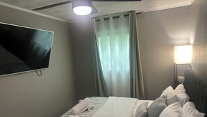 A neatly made bed with white linens and pillows, a flat-screen TV mounted on the wall, a bedside lamp, and a window with sheer curtains.