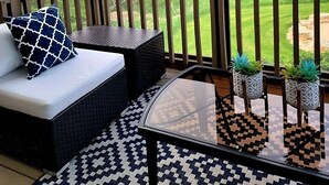 Un espacio de estar al aire libre cubierto, con un sofá, cojines y una mesa de vidrio decorada con plantas en macetas.