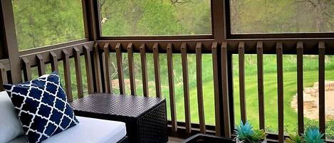 Un espacio de estar al aire libre cubierto, con un sofá, cojines y una mesa de vidrio decorada con plantas en macetas.
