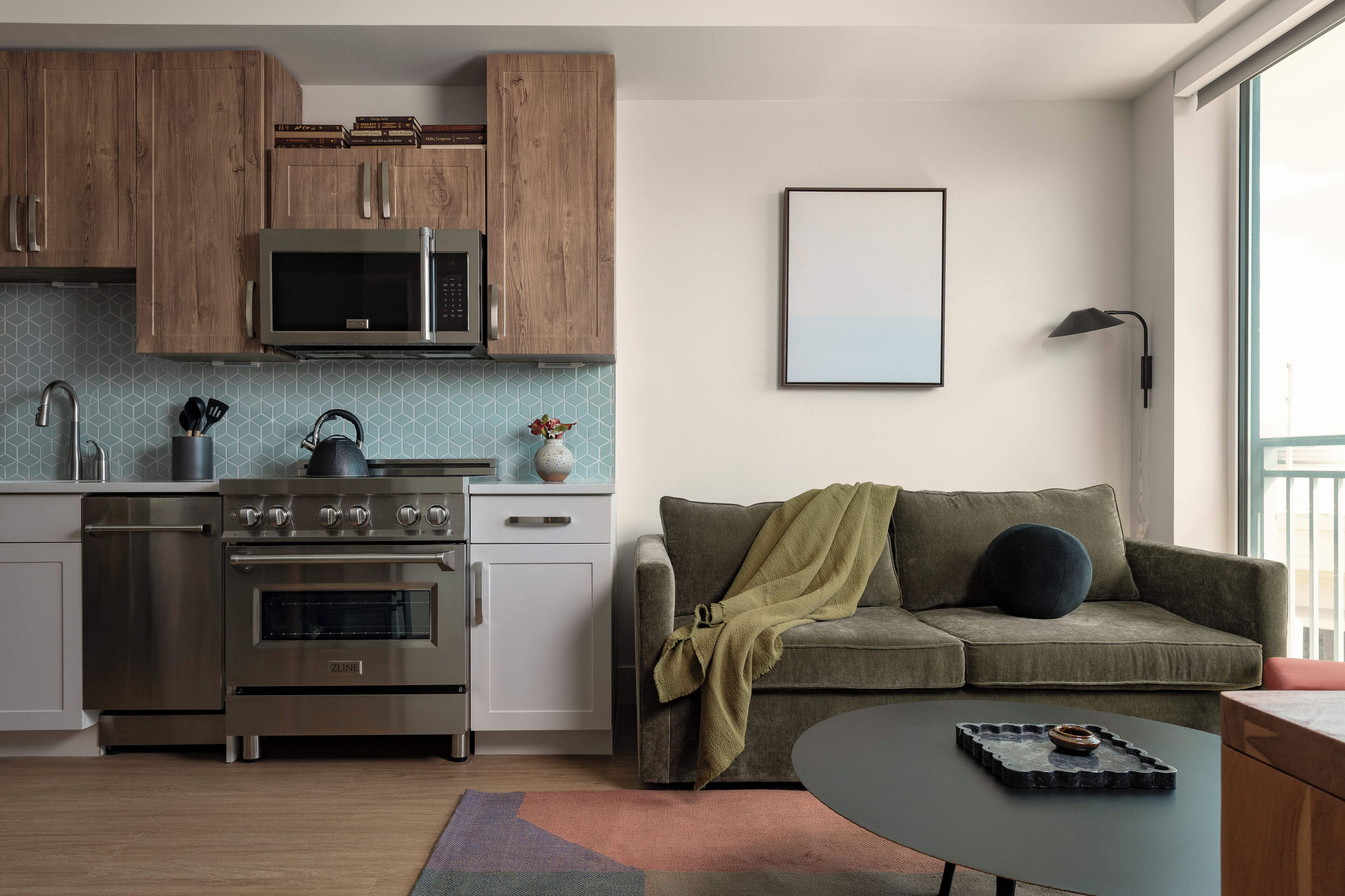 A modern kitchen with wooden cabinets, a stainless steel stove, and a microwave. There is a green sofa with a blanket and a black lamp on the wall.