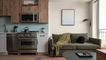 A modern kitchen with wooden cabinets, a stainless steel stove, and a microwave. There is a green sofa with a blanket and a black lamp on the wall.