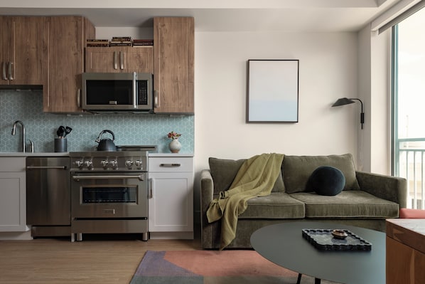 Une cuisine moderne avec des meubles en bois, une cuisinière en acier inoxydable et un four à micro-ondes. On y trouve un canapé vert avec une couverture et une lampe noire fixée au mur.