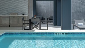 Un área de piscina interior moderna con borde de azulejos, una mesa con sillas y un espacio de descanso con sillones.