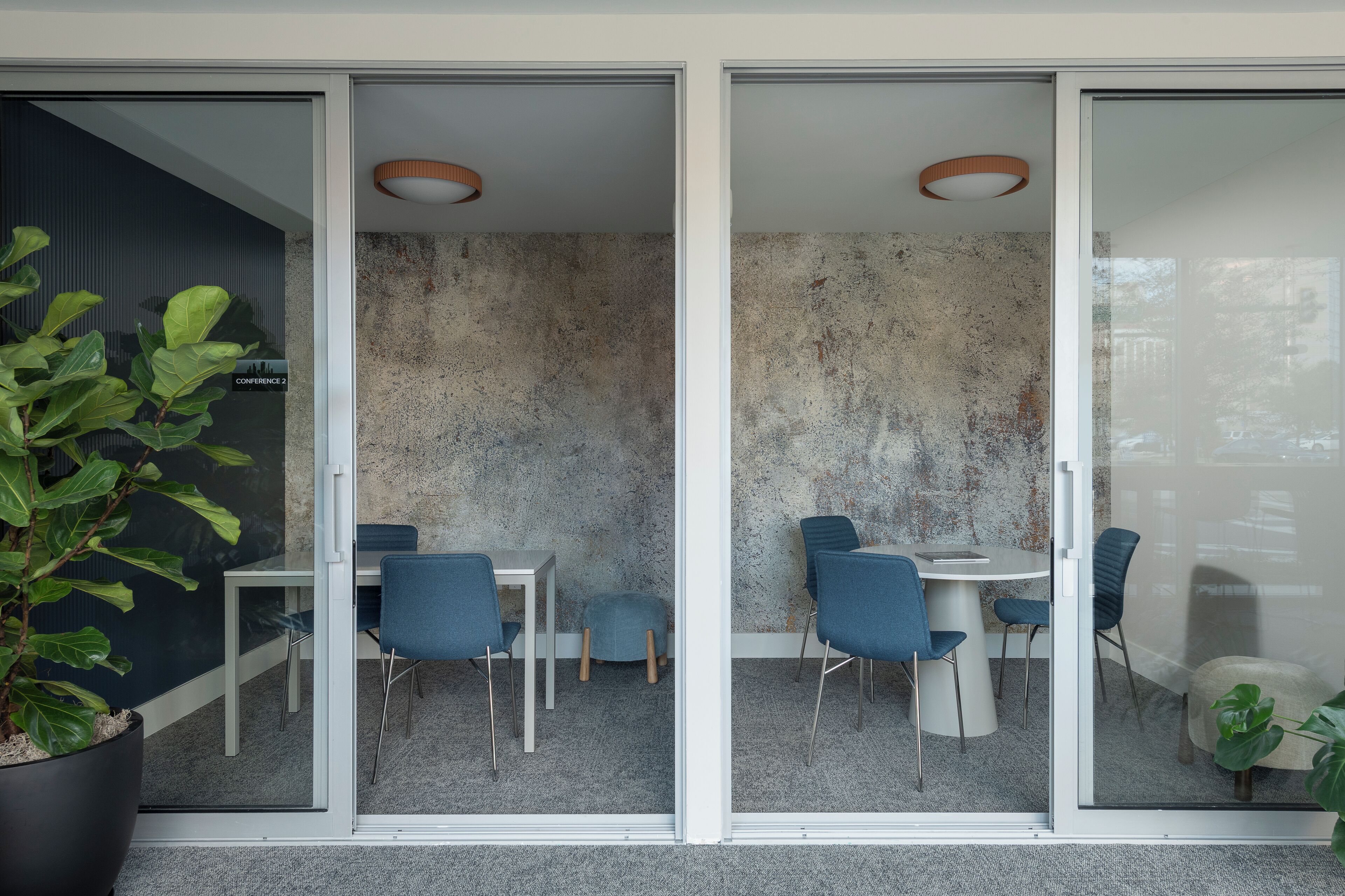 Uma sala de reuniões moderna com paredes de vidro, mesa branca, cadeiras azuis e um vaso de planta.