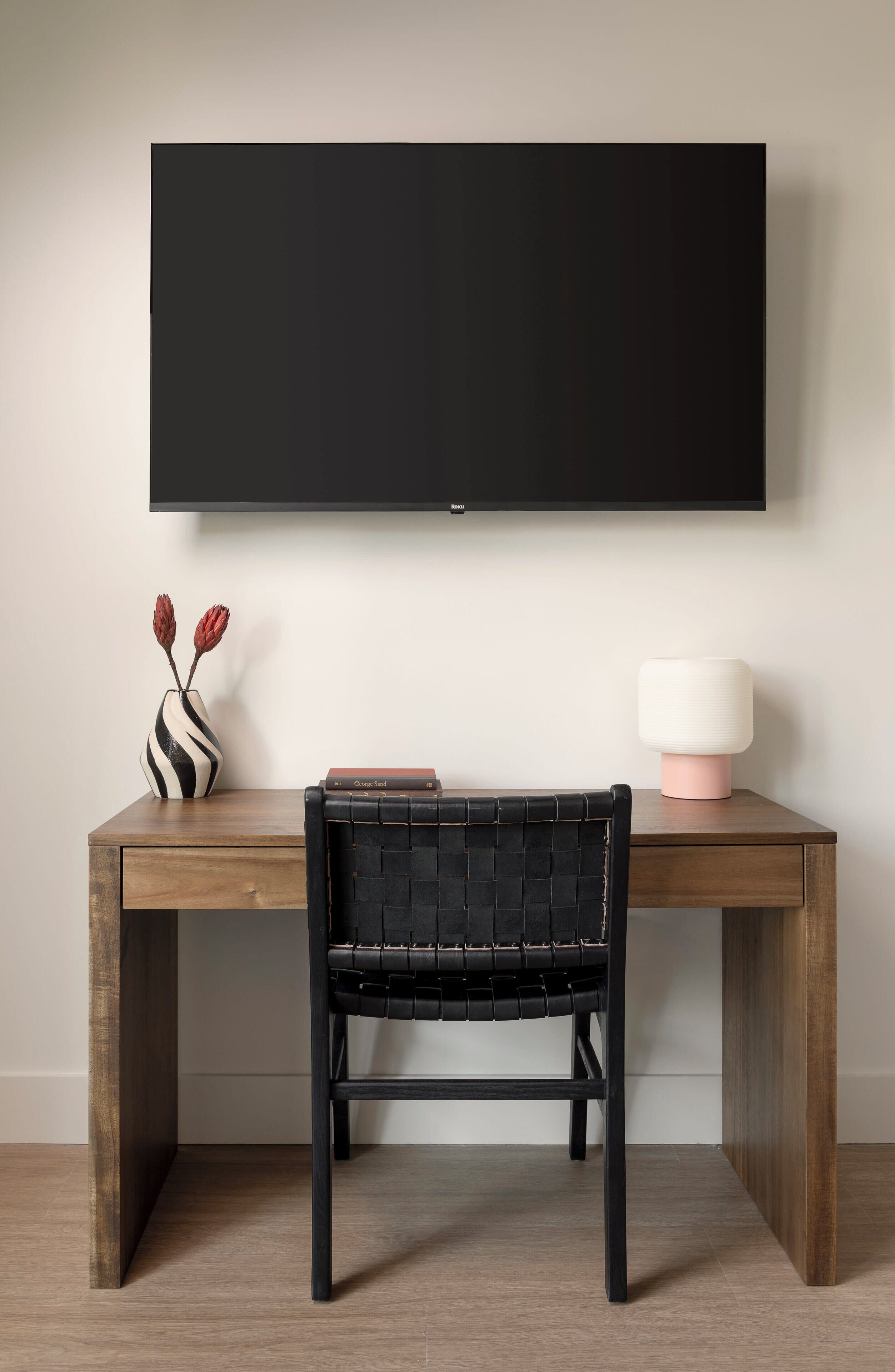 A wooden desk with a black woven chair, a black flat-screen TV mounted above, a striped vase with red flowers, and a pink lamp on the right side.