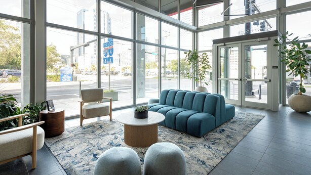 Un moderno vestíbulo de hotel con ventanas amplias, un sofá azul, una mesa de centro redonda y una zona de estar con una alfombra pequeña.