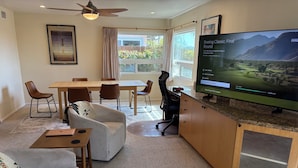 Una sala de estar moderna con un televisor de pantalla plana grande mostrando un torneo de golf, una zona de comedor con una mesa y sillas de madera, un sofá y un ventilador de techo.