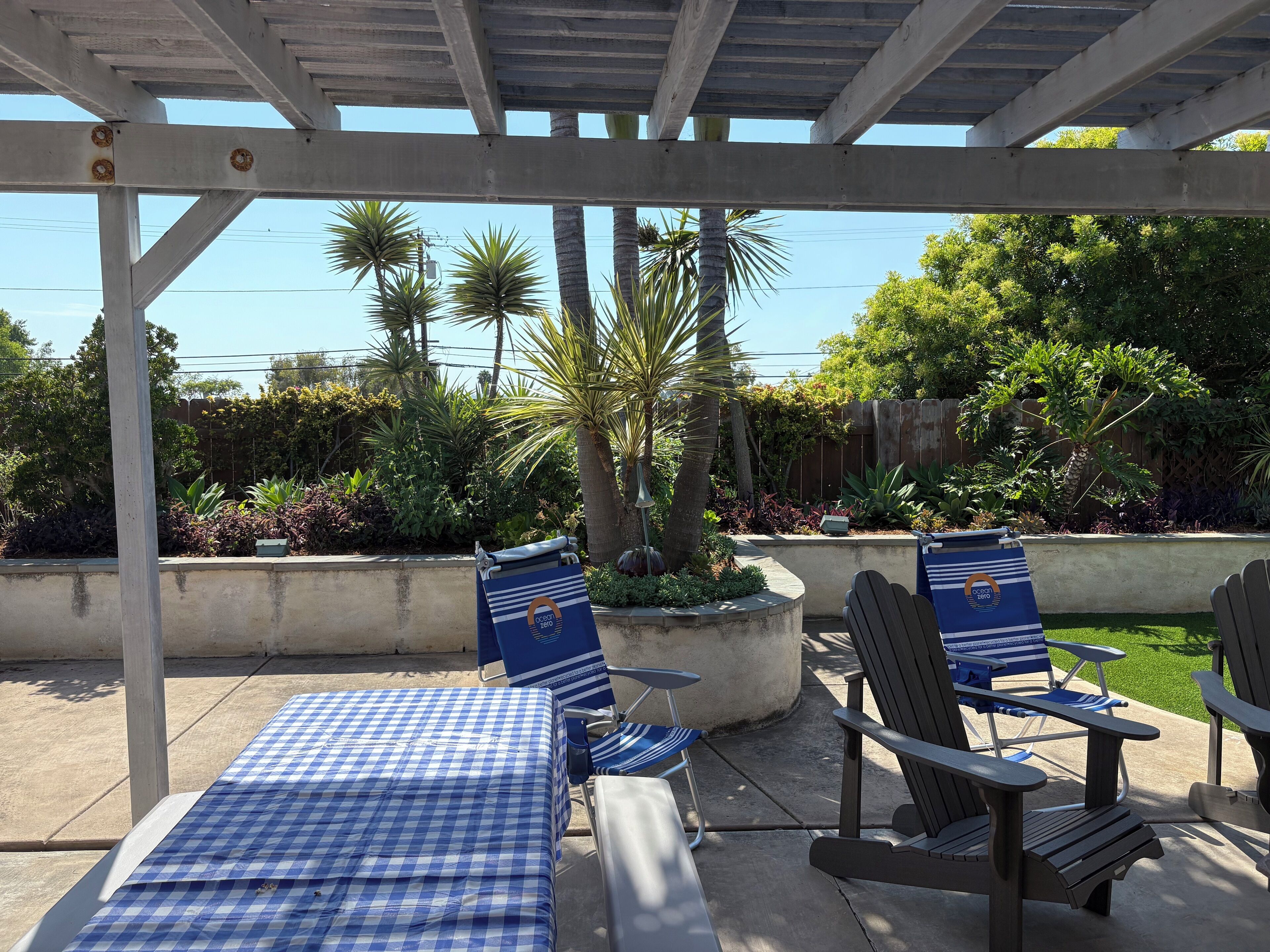 A covered patio area with checkered tablecloths, Adirondack chairs, and a concrete planter with a palm tree.