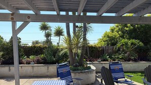 Un patio cubierto con manteles a cuadros, sillas Adirondack y una jardinera de concreto con una palmera.