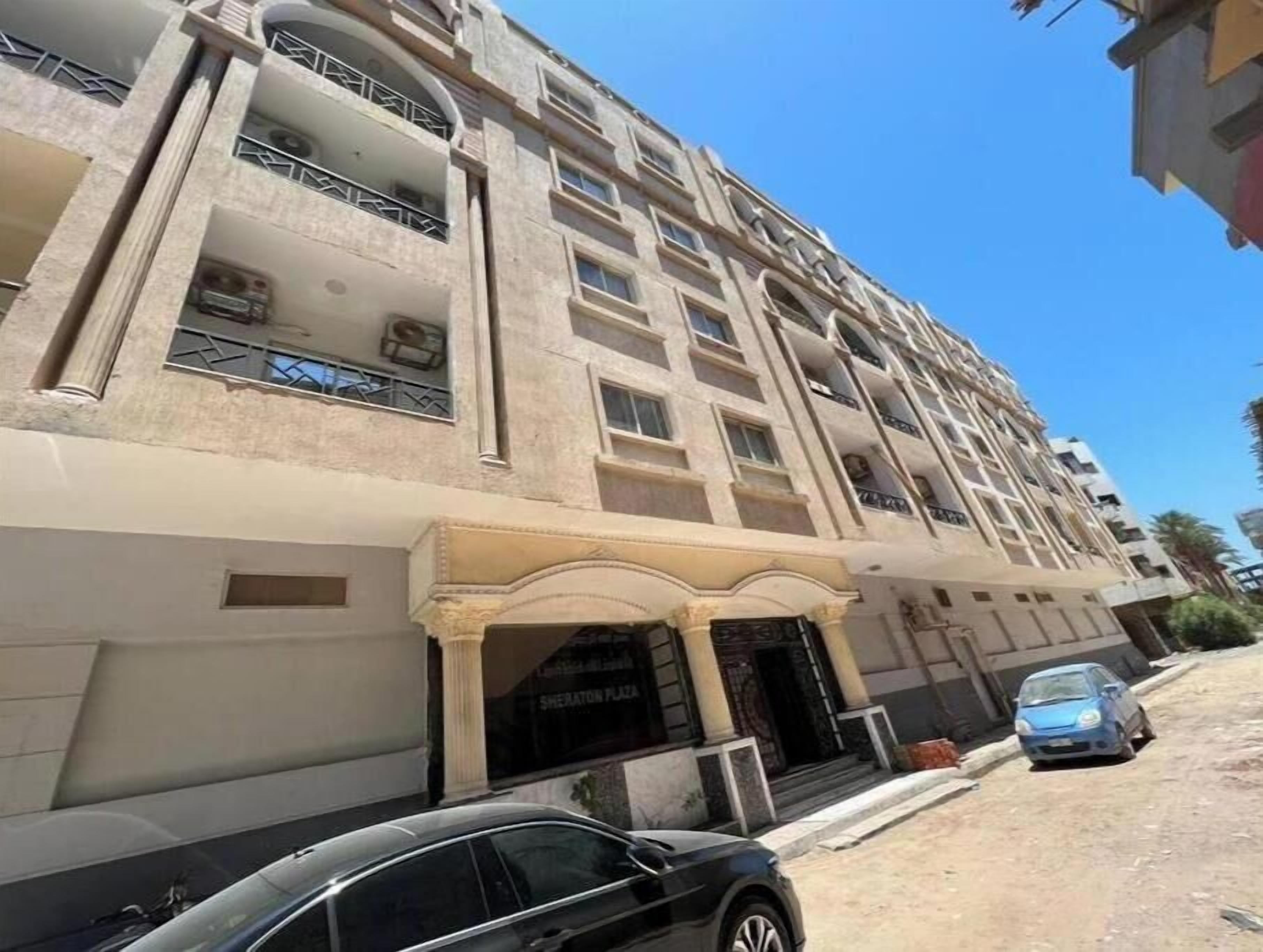 A multi-story building with a central entrance, balconies, and a parking area with cars.