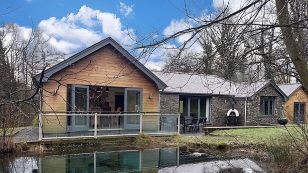 A house with a wooden exterior, a glass door entrance, a small outdoor seating area, and a pond in the foreground. - Bracken Ground (Ulverston)