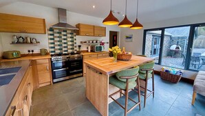 A modern kitchen with wooden cabinets, a central island, bar stools, and a view to the outside. - Bracken Ground (Ulverston)