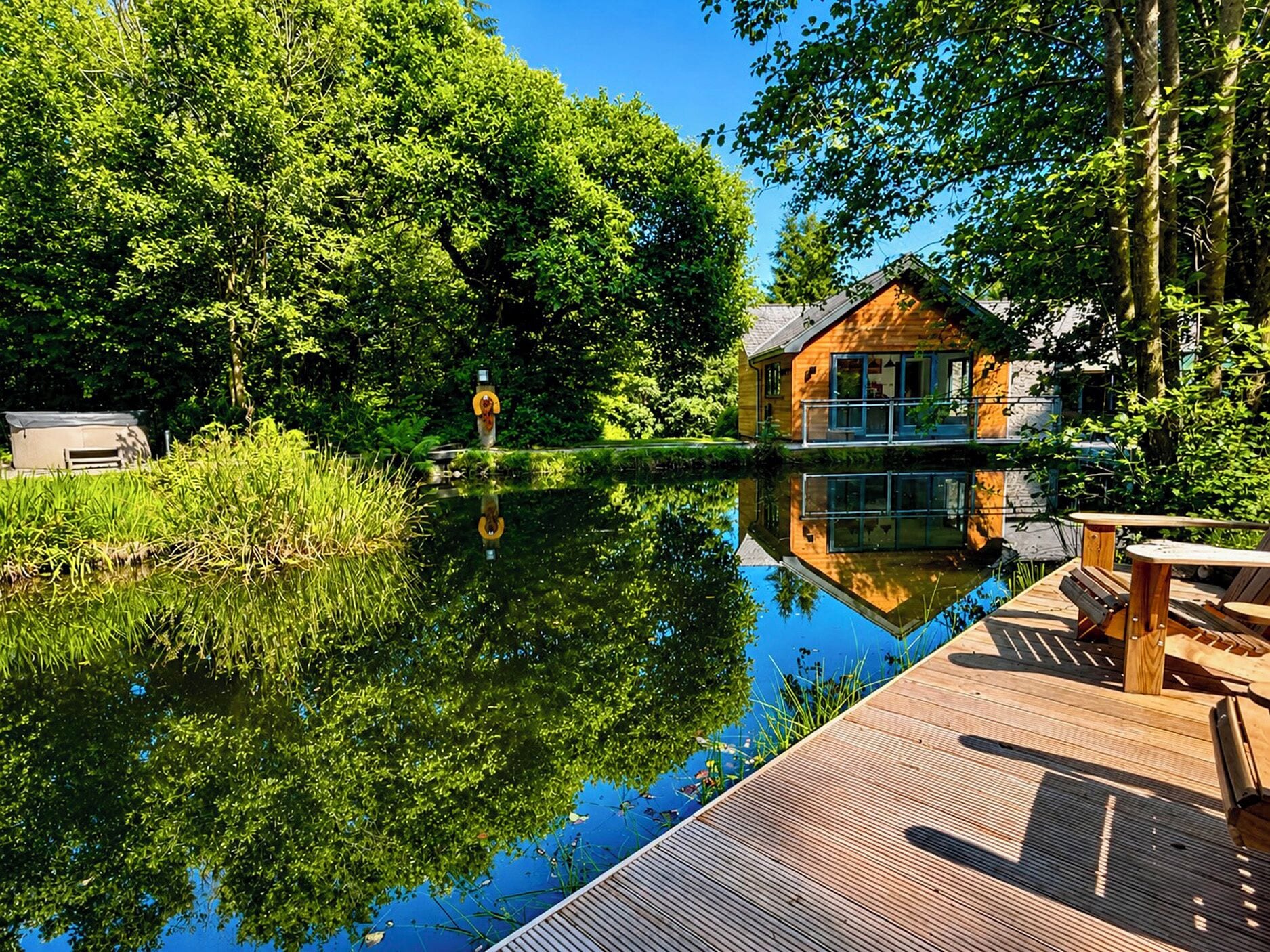 A wooden deck with a view of a house across a calm lake, surrounded by lush greenery.