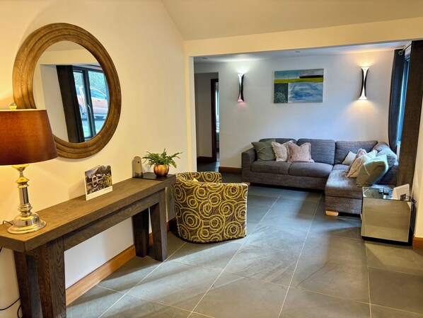 A living room with a wooden console table, a round mirror, a patterned ottoman, a grey sofa with cushions, and a painting on the wall. - Bracken Ground (Ulverston)
