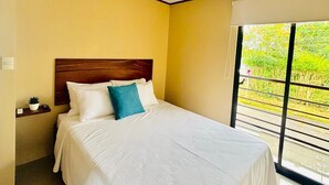 Habitación de hotel con una cama grande, cabecera de madera, mesita de noche pequeña con una planta y ventana con vistas a la vegetación.