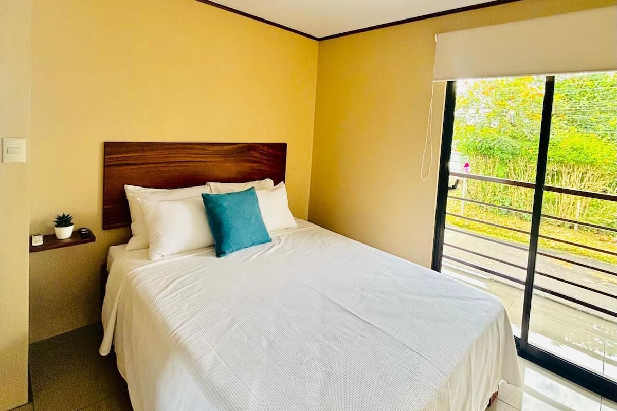 Une chambre d’hôtel avec un grand lit, une tête de lit en bois, une petite table de chevet agrémentée d’une plante et une fenêtre donnant sur un paysage verdoyant.