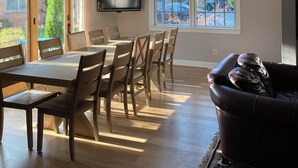 A spacious dining area with a large table and chairs, a flat-screen TV mounted on the wall, and large windows offering views of the outdoors.