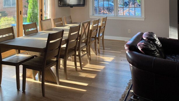 A spacious dining area with a large table and chairs, a flat-screen TV mounted on the wall, and large windows offering views of the outdoors.