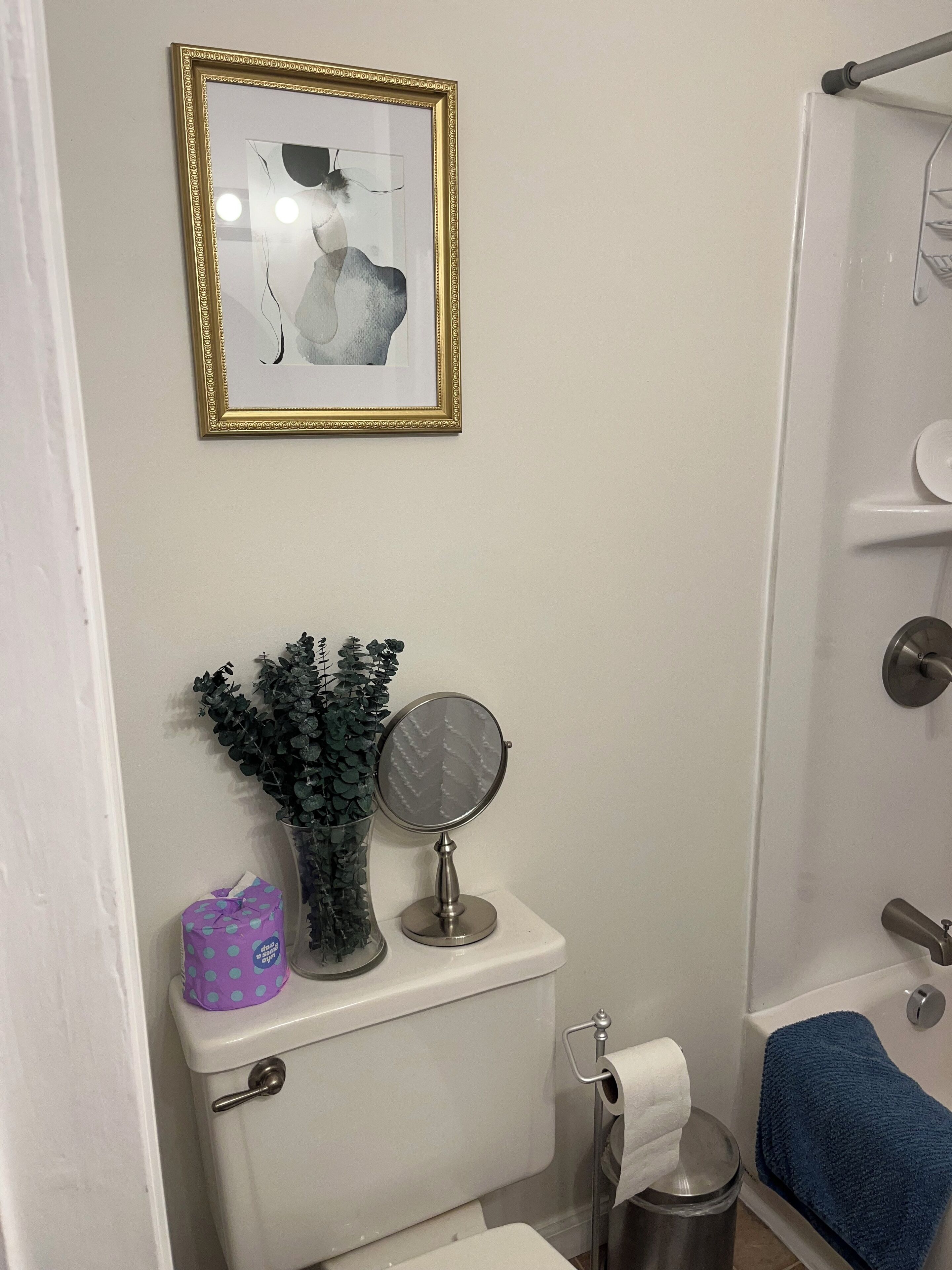 Ein Badezimmer mit Toilette, Waschbecken, Dusche, einem gerahmten Kunstwerk, einer Zimmerpflanze und einem Spiegel.
