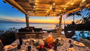 A dining table set for two with a view of the ocean at sunset, under a wooden pergola with hanging lanterns. - Artist Cliffside Home with private beach access and cabana and two cats. (Punta de mita Bahía de banderas)