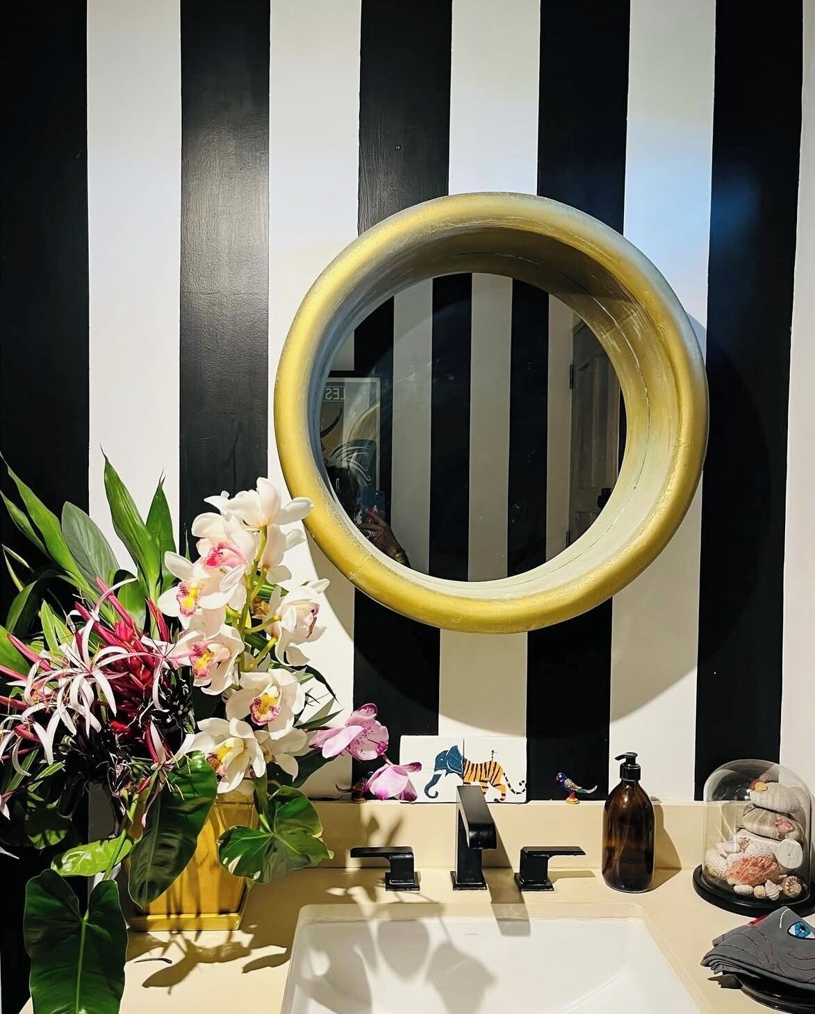 Een badkamer met een zwart-wit gestreepte wand, een ronde spiegel, een wastafel, een toilet en een vaas met bloemen.