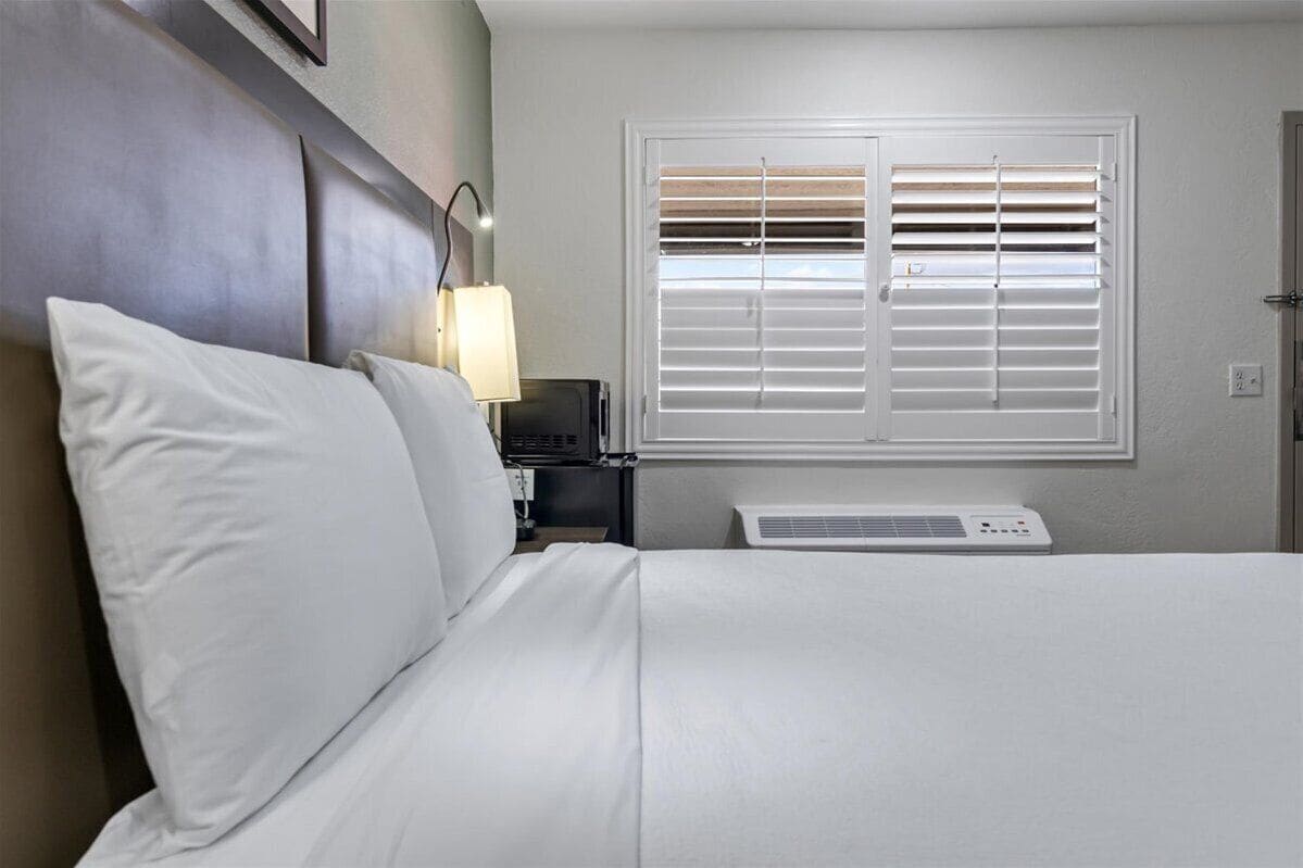 Una habitación de hotel con cama, mesita de noche con lámpara, ventana con persianas y un equipo de aire acondicionado.