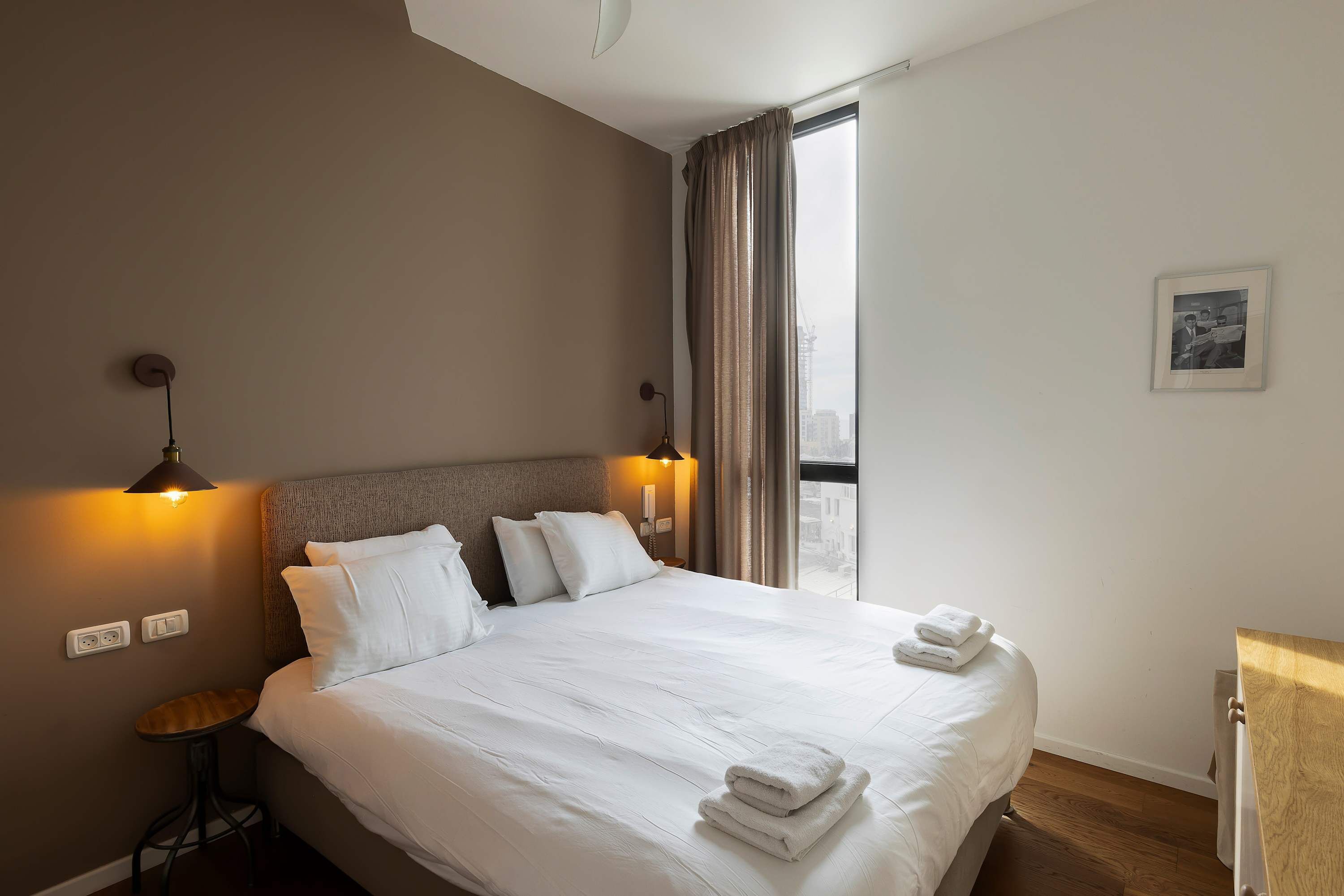 Ein ordentlich gemachtes Bett mit weißen Leinen und Handtüchern, ein Nachttisch mit Lampe und ein Fenster mit Vorhängen.