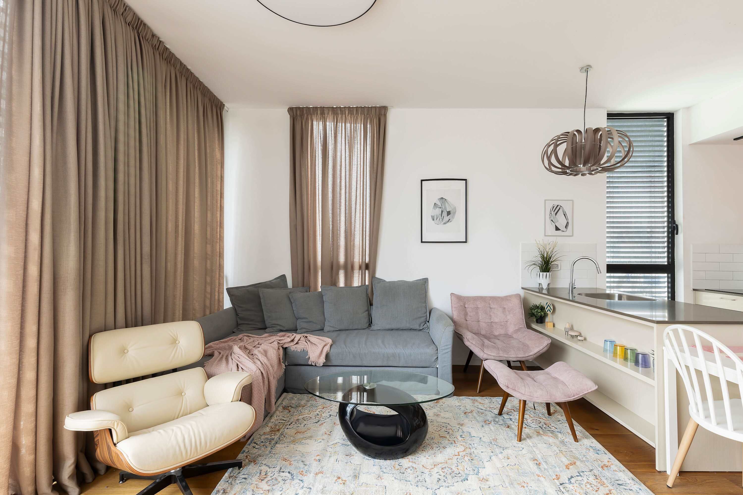 A modern living room with a grey sofa, a beige armchair, a glass coffee table, and a white chair. A kitchen area is visible to the right with a white countertop and a hanging light fixture.