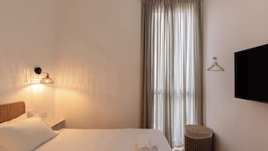 A hotel room with a bed, a wall-mounted TV, a bedside lamp, a window with curtains, and a small round stool. - Stylish Beach Getaway  Steps from Carmel Market! (Tel Aviv-Yafo)