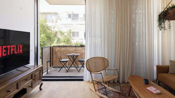 A modern living room with a flat-screen TV, a sofa, a coffee table, and a balcony with a table and chairs. - Stylish Beach Getaway  Steps from Carmel Market! (Tel Aviv-Yafo)