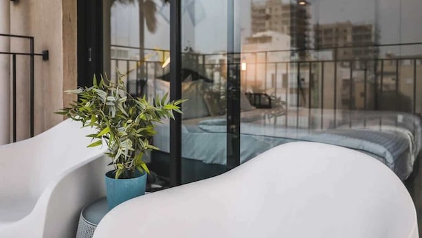 A modern balcony with a white sofa and a potted plant, overlooking a cityscape with buildings and a construction crane.