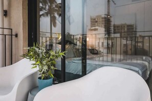 Un balcón moderno con un sofá blanco y una planta en maceta, con vistas a un paisaje urbano con edificios y una grúa de construcción.