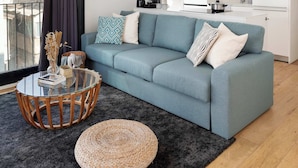 A modern living room with a blue sofa, a glass coffee table, and a woven pouf. A kitchen with white cabinets and a black countertop is visible in the background.