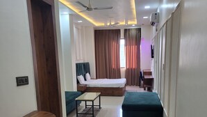 A modern hotel room with a bed, a desk, a chair, a sofa, and a TV. - Hotel MG Kitchen and Banquet (Kathua)