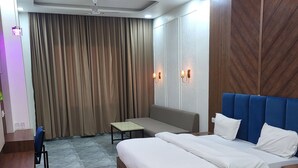 A modern hotel room with a wooden floor, a large bed with white bedding, a blue headboard, a desk, a chair, a small table, and a ceiling fan. - Hotel MG Kitchen and Banquet (Kathua)