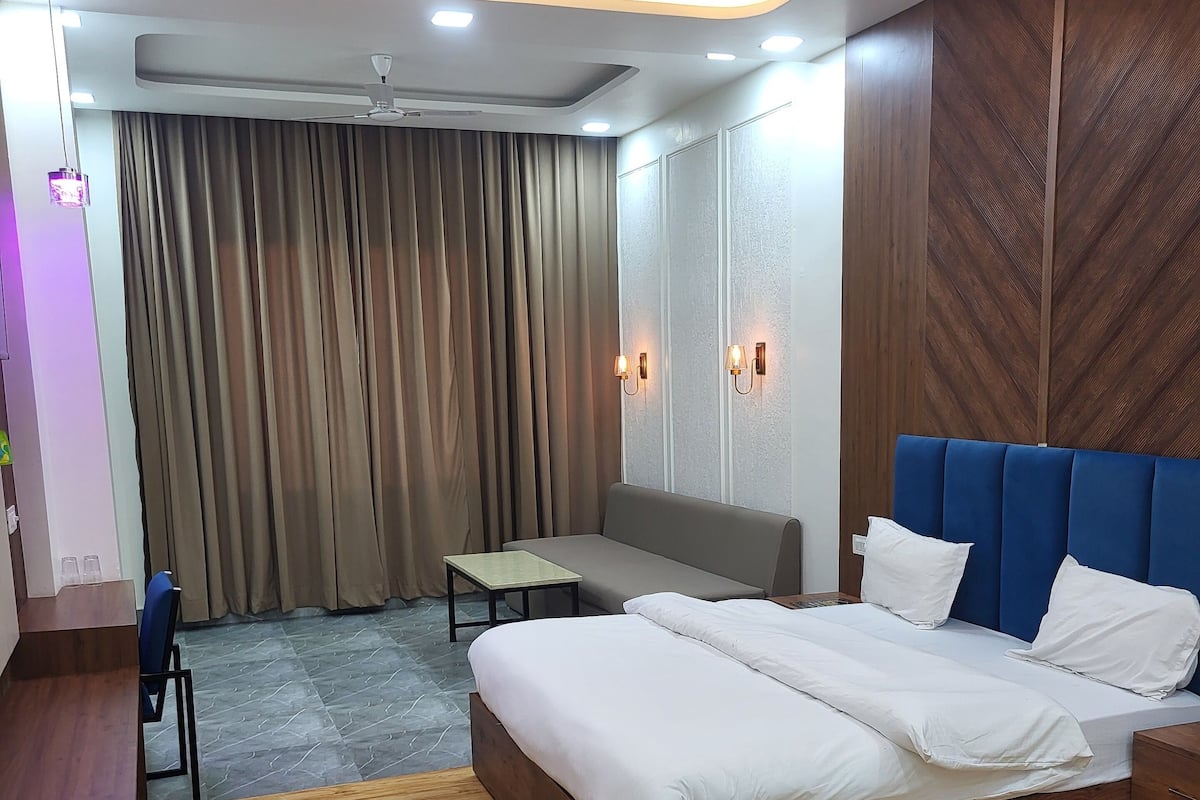 Una habitación de hotel moderna con piso de madera, una cama grande con ropa blanca, un cabecero azul, un escritorio, una silla, una mesita y un ventilador de techo.