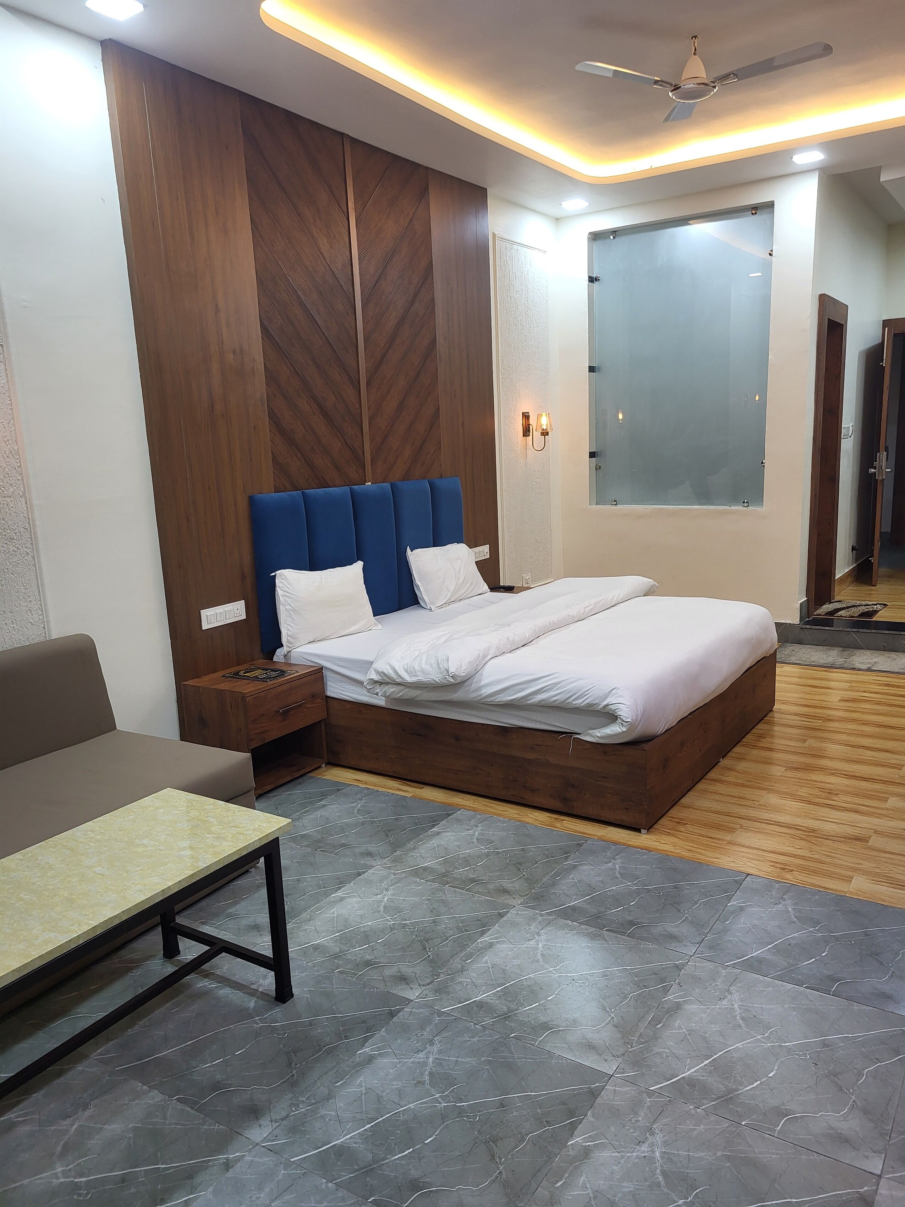 Un dormitorio moderno con una cama grande, cabecera de madera, mesita de noche, zona de estar y un amplio ventanal.