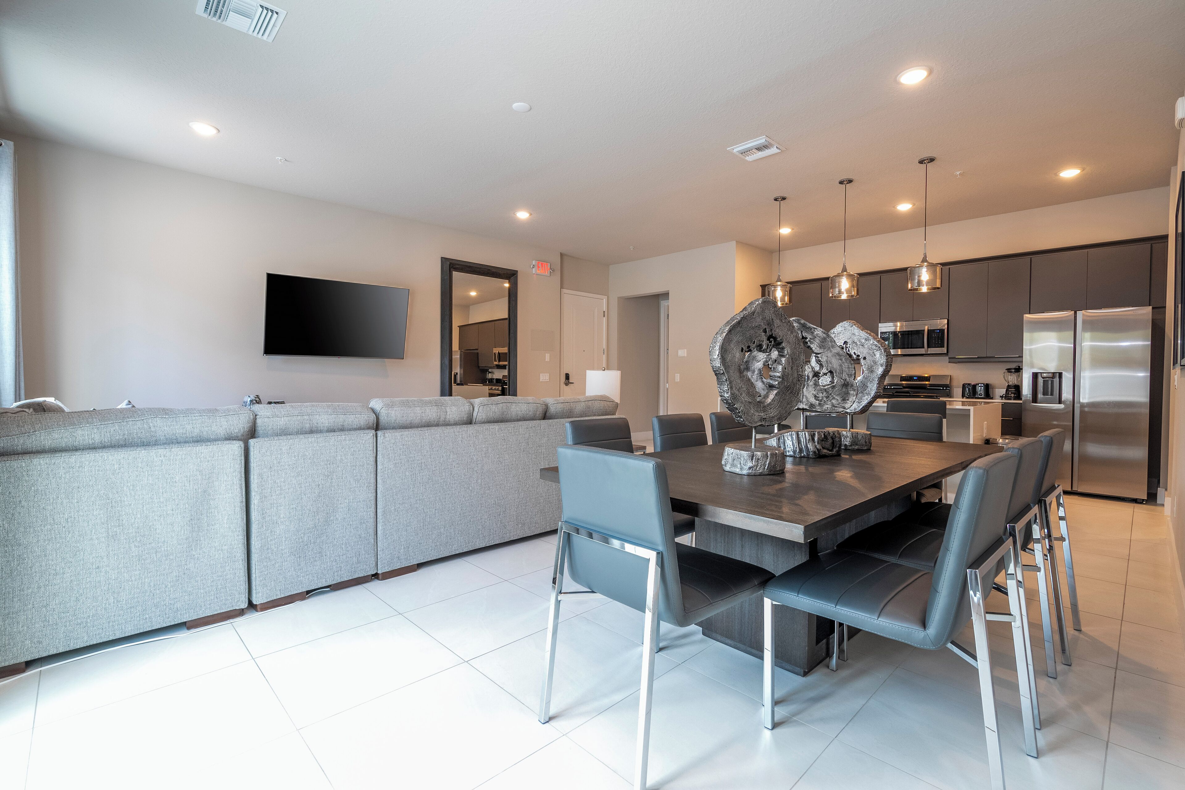 A modern kitchen with stainless steel appliances, a dining area with a large table and chairs, and a grey sofa in the living area.