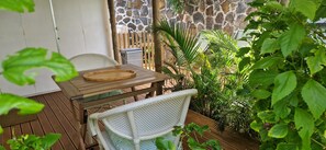 An outdoor wooden deck with a table and chairs, surrounded by lush greenery and a stone wall.