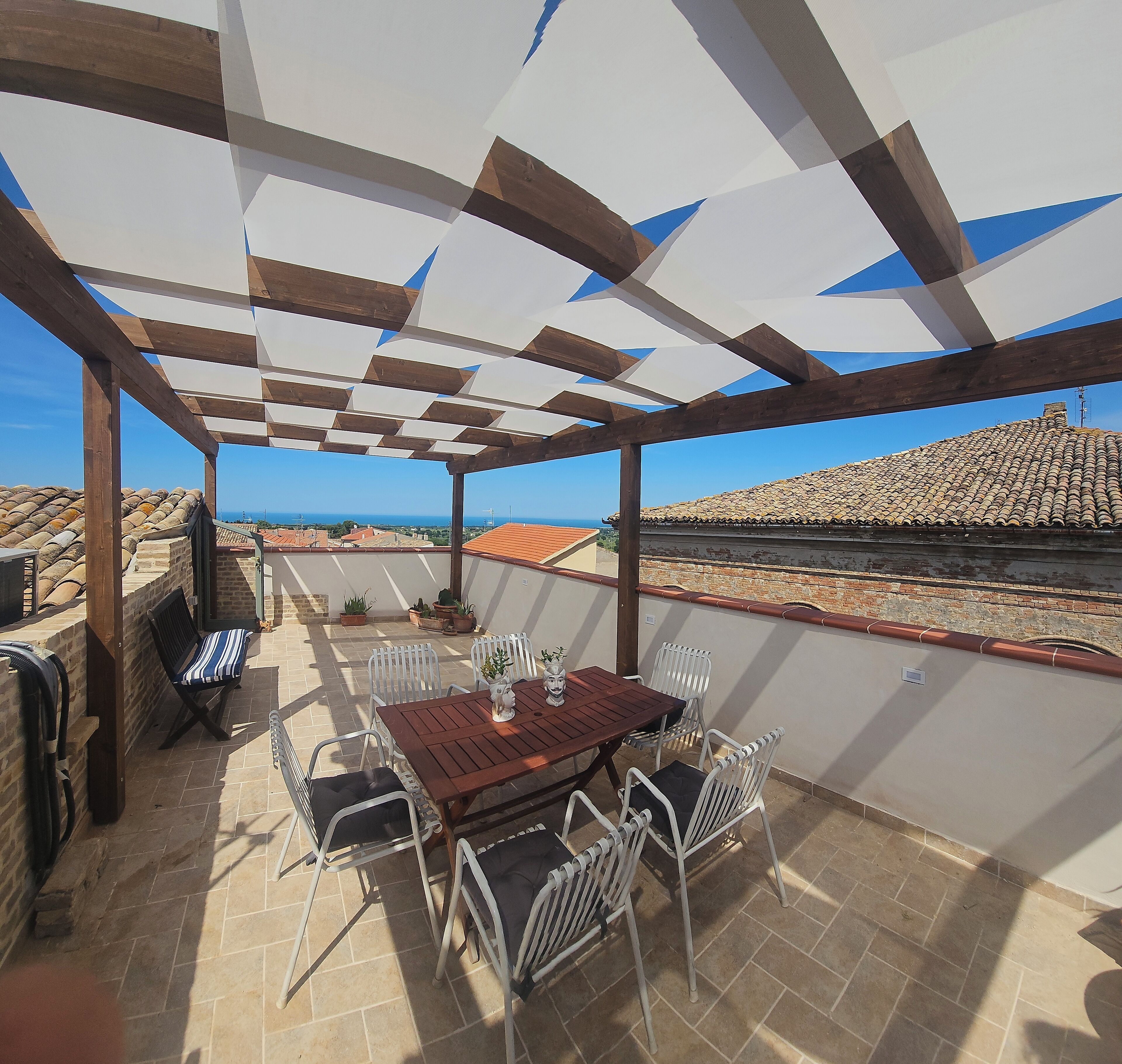 A covered patio with a wooden table and chairs, potted plants, and a view of the sea and buildings in the background.