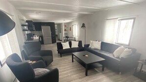 Ein modernes Wohnzimmer mit einer Couch, Sesseln und einem Couchtisch. Im Hintergrund ist eine Küchenzeile mit einem Kühlschrank und Schränken zu sehen.