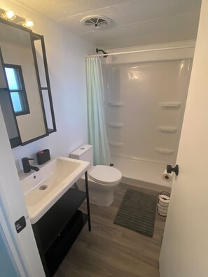 Un baño con lavamanos, inodoro y ducha blancos. Hay un espejo sobre el lavamanos y un perchero encima del inodoro.