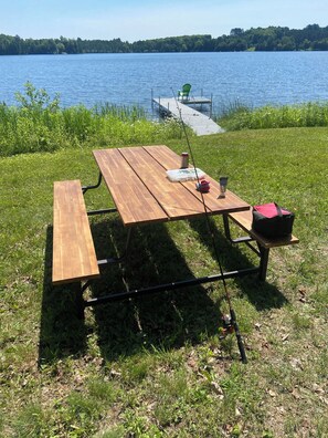 Una mesa de picnic de madera con bancos, una caña de pescar y una bolsa roja en un césped cerca de un lago.