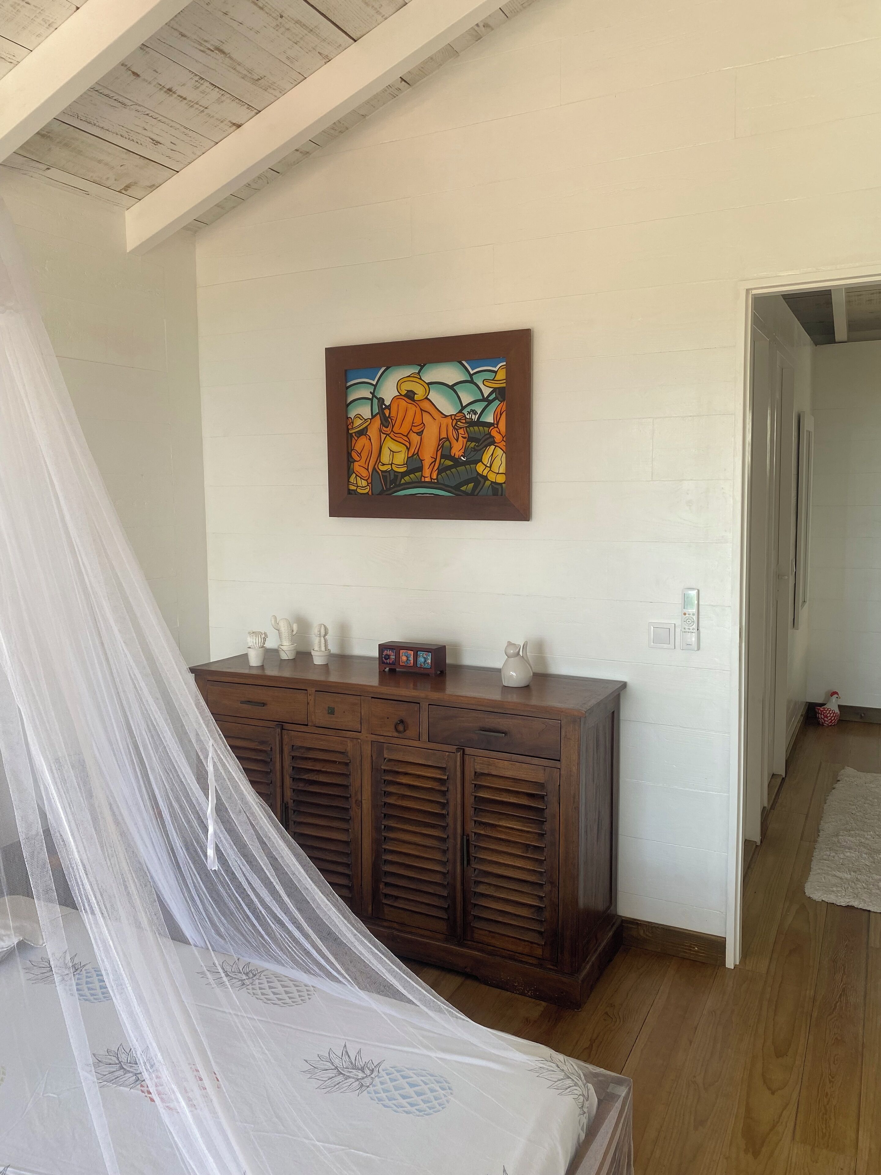 A bedroom with a bed covered by a mosquito net, a wooden cabinet with decorative items, and a painting on the wall.