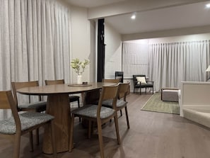 A dining area with a round wooden table and six chairs, a small side table with a vase, and a view of a living room with a sofa and a chair. - Chalet 6 @ The Retreat (Taupo)