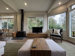 Ein modernes Wohnzimmer mit Kamin, Fernseher und großen Fenstern mit Blick auf Grünflächen.