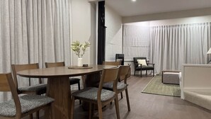 Un comedor con una mesa redonda de madera y seis sillas, una mesita auxiliar con un jarrón, y vistas a una sala de estar con un sofá y una butaca.