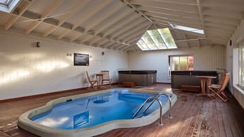An indoor swimming pool with a wooden deck, a hot tub, and seating areas.