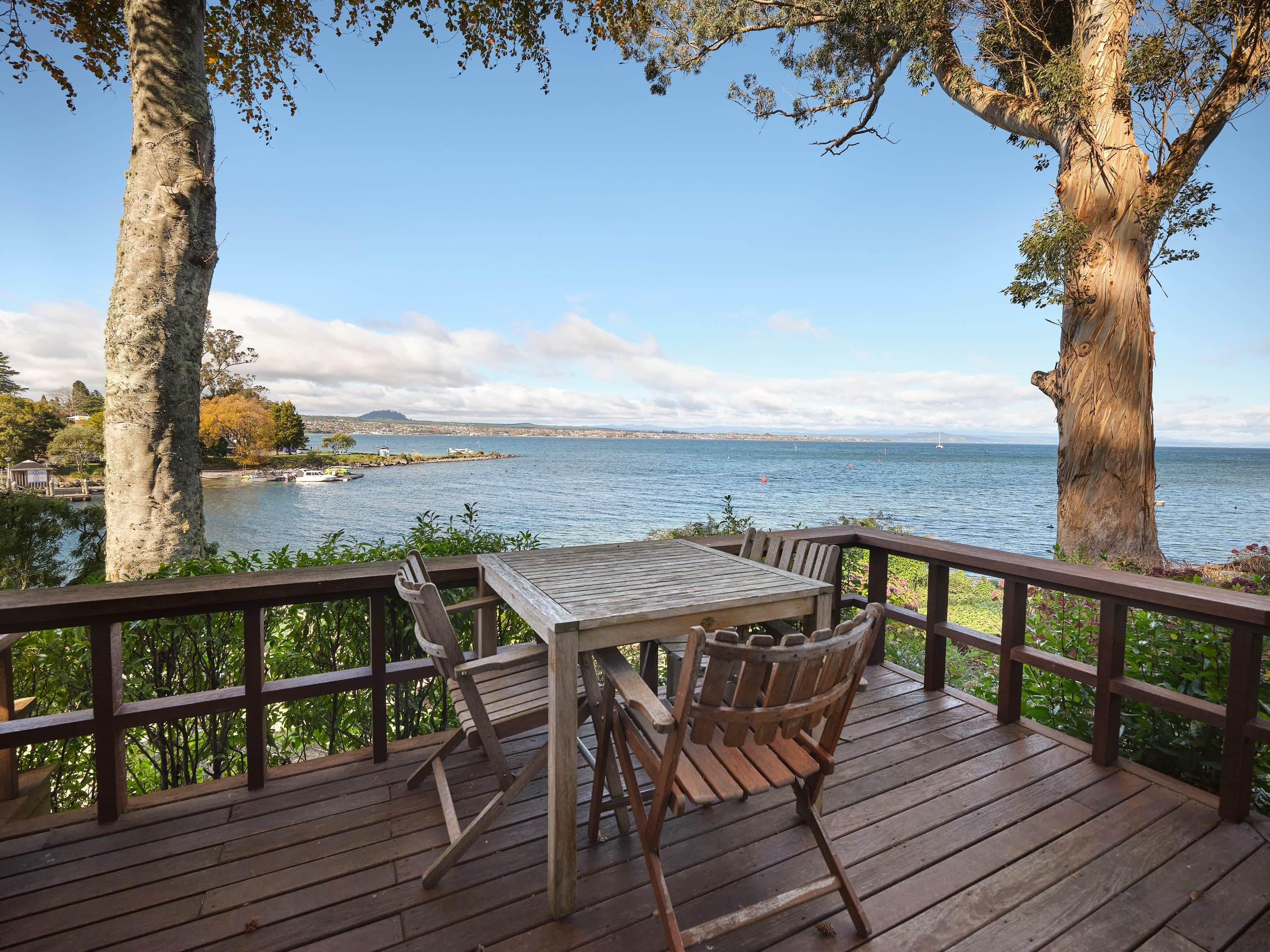 A wooden deck with a table and chairs overlooking a body of water with boats and a distant shoreline.